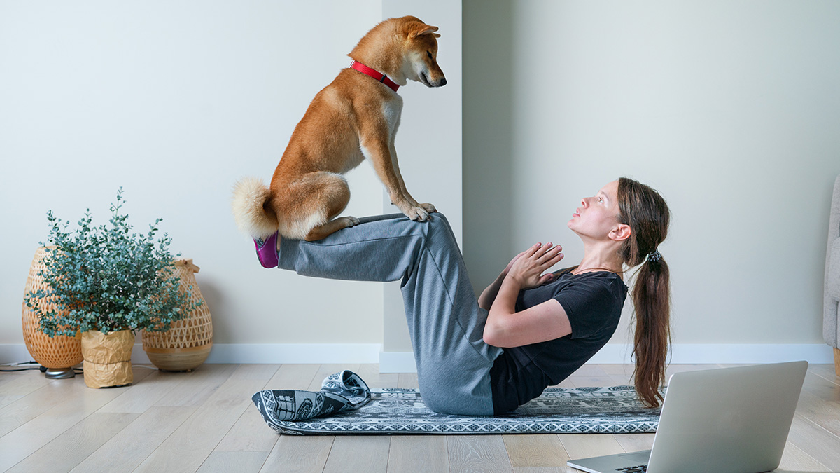 A young woman doing yoga with her dog, lifting up her legs to strengthen the core, a dog sitting on her shins